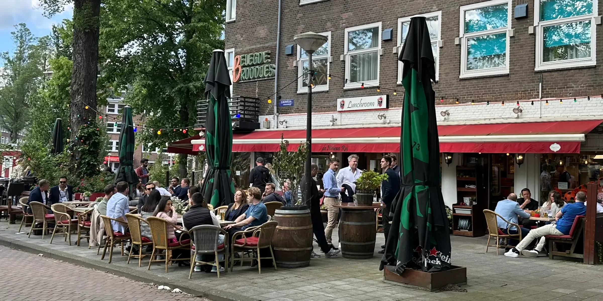 Druk terras met gasten onder de rode luifels van Broodjeszaak De Nieuwe Rai in Amsterdam Zuid